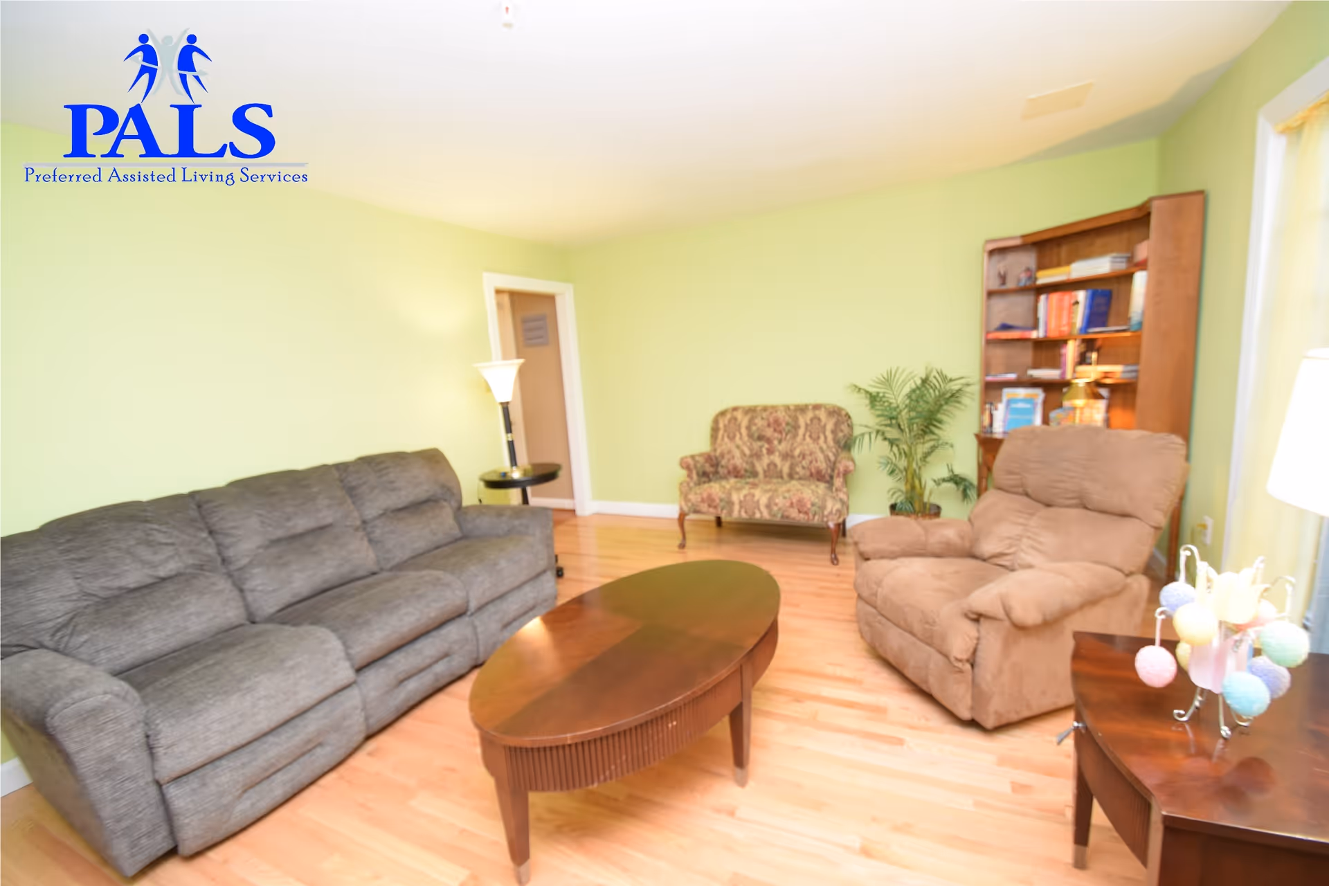 A cozy living room with light green walls and hardwood floors. The room features a gray three-seater sofa, a brown recliner chair, a floral-patterned loveseat, a wooden oval coffee table, a wooden bookshelf filled with books, a potted plant, and two side tables with lamps. The logo for Preferred Assisted Living Services (PALS) is visible in the top left corner.