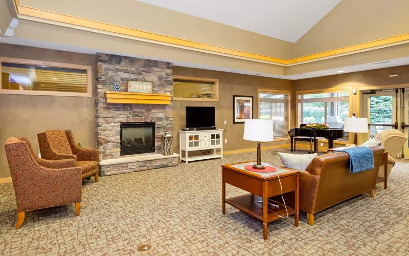 A cozy living room area in an assisted living facility featuring a stone fireplace with a wooden mantel displaying a 'SWEET' sign, two patterned armchairs, a brown leather sofa with a blue throw, a wooden side table with a lamp, a white TV stand with a flat-screen TV, large windows with blinds, and a black grand piano near the entrance door.