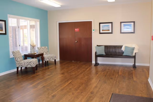 A room with wooden flooring featuring two patterned armchairs and a small wooden table near a window on the left side, and a wooden bench with two blue cushions and a white throw on the right side. The back wall has two framed pictures and a double wooden door with a red 'NO EXIT' sign.