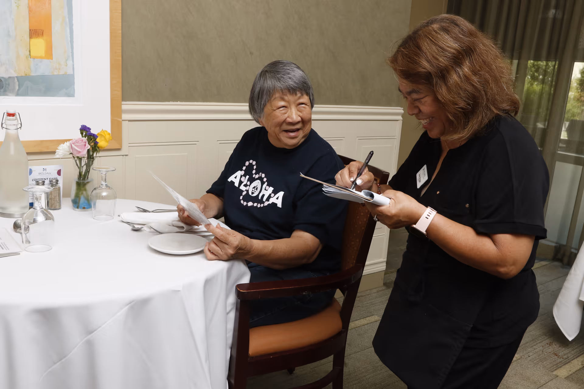 An elderly woman wearing a black Aloha t-shirt is seated at a dining table with a white tablecloth, holding a menu and smiling at a waitress who is standing beside her taking notes on a pad. The table has a small vase with flowers, glassware, and a water bottle. The setting appears to be a dining room with light-colored walls and a framed artwork.