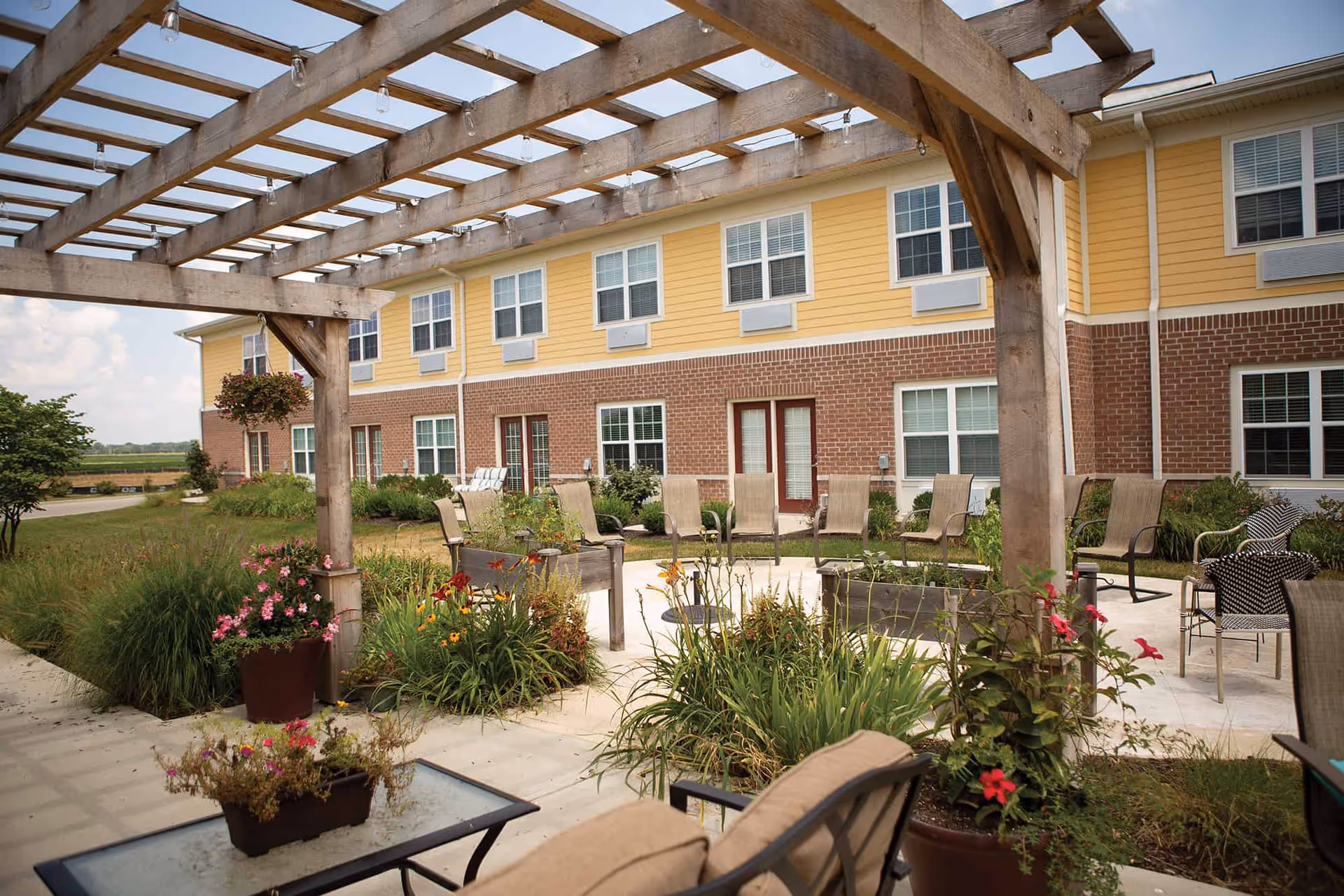 Outdoor patio area at Traditions at Reagan Park featuring a wooden pergola, multiple chairs arranged in a circle around a fire pit, potted plants with flowers, and a two-story building with yellow siding and brick exterior in the background.