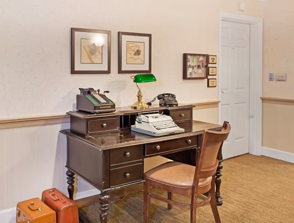 A vintage wooden desk with a green banker’s lamp, an old typewriter, a rotary telephone, and a adding machine on top. Two framed pictures hang on the wall above the desk. A wooden chair is placed in front of the desk. To the right, there is a white door and a bulletin board with room number 119 and name tags. Two vintage suitcases are on the floor beside the desk. The room has beige wallpaper and carpeted floor.