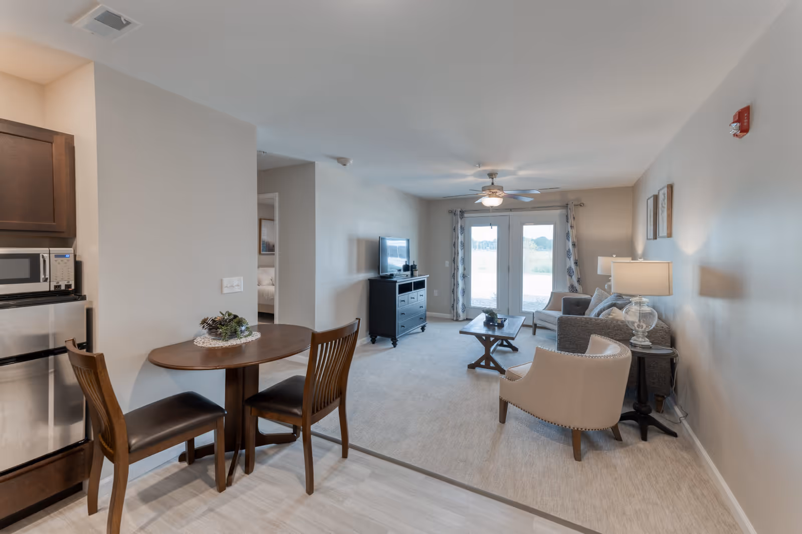 A senior living facility interior showing a small dining area with a round wooden table and two chairs next to a kitchenette with a microwave and mini fridge. The living room area features a sofa, armchair, coffee table, TV on a stand, and glass doors leading outside. The room is decorated in neutral tones with a ceiling fan and lamps providing lighting.