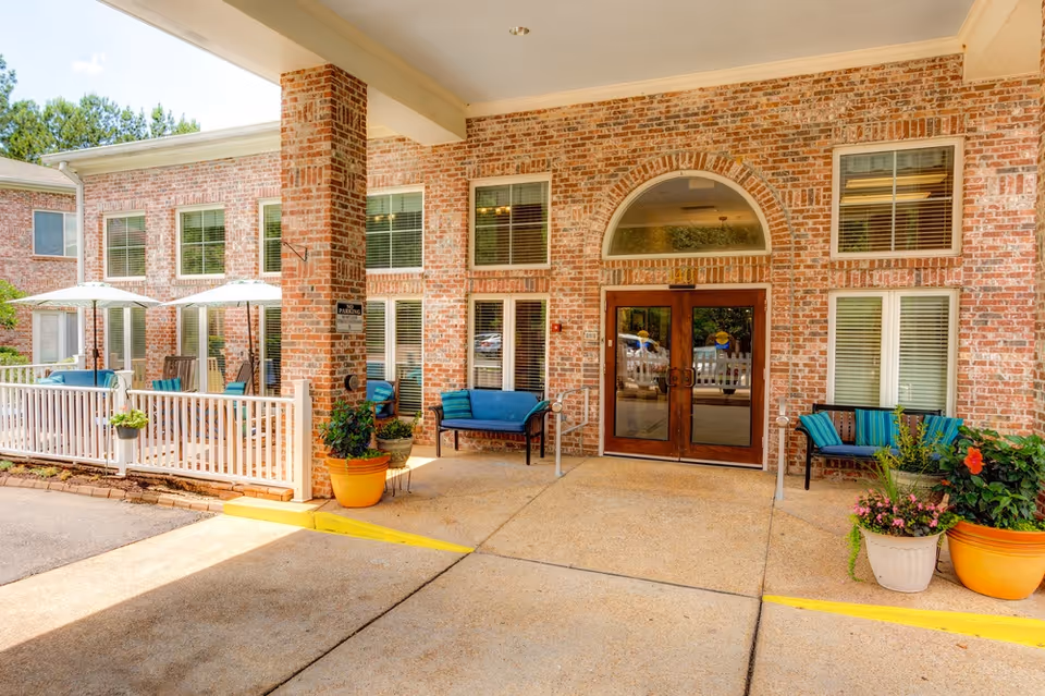 Covered main entrance of a brick senior living facility with double glass doors, outdoor seating, umbrellas and potted plants.