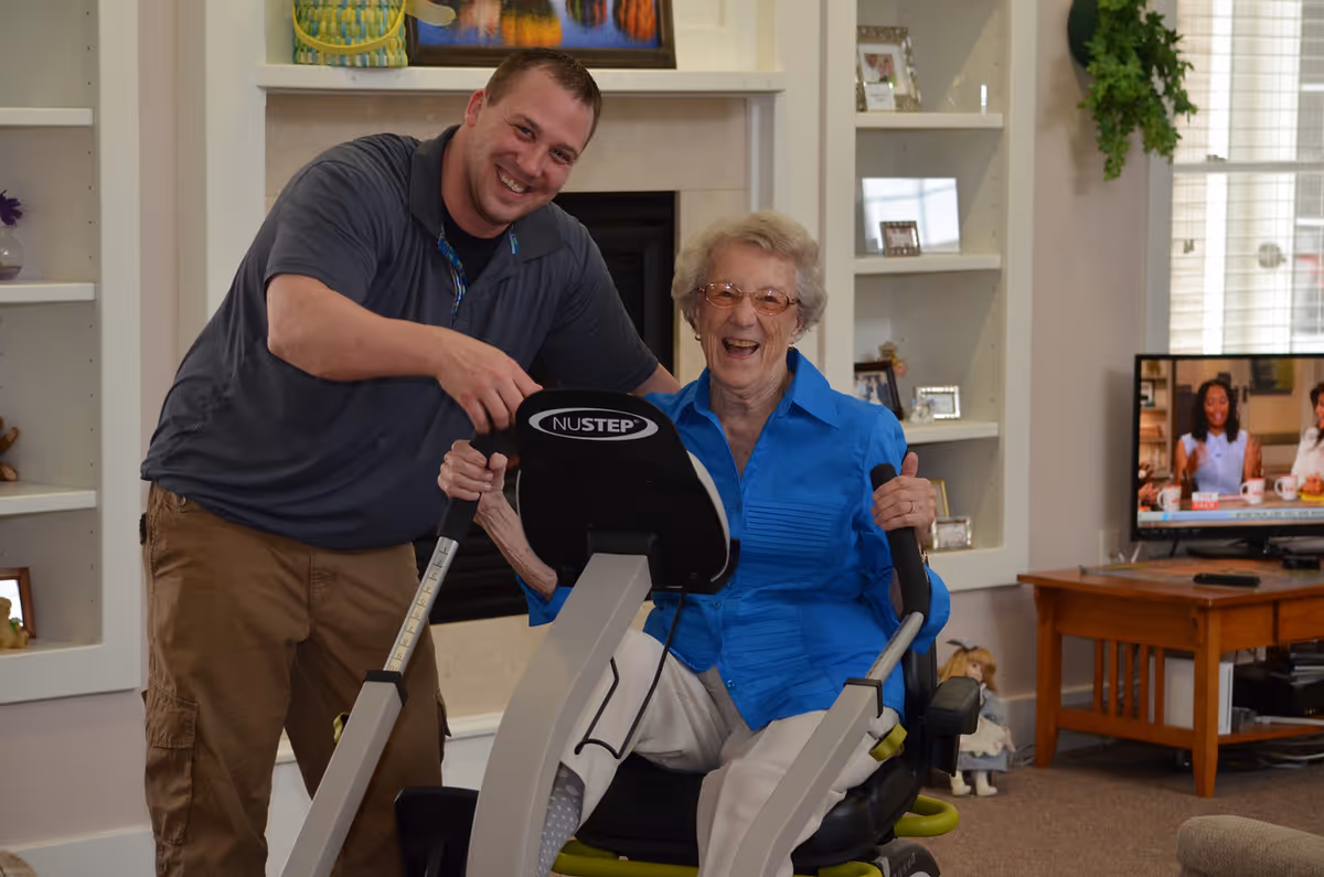 An elderly woman wearing a blue shirt is sitting on a NuStep exercise machine, smiling and holding the handles. A man in a gray shirt and khaki pants is standing next to her, also smiling and assisting with the exercise. They are in a cozy living room with built-in shelves, a fireplace, and a television showing a talk show in the background.