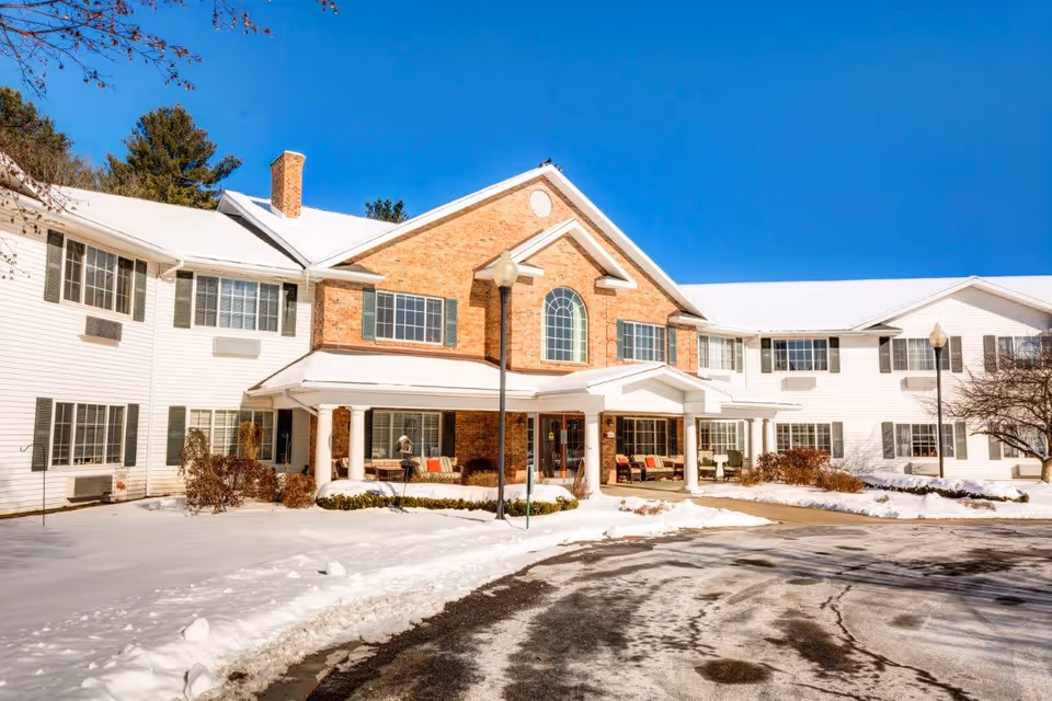 Exterior view of The Landing at Queensbury senior living facility on a sunny winter day with snow covering the ground and roof. The building features white siding with green shutters and a central brick section with large windows and a covered entrance with columns. There are outdoor seating areas on the porch and a curved driveway in front.