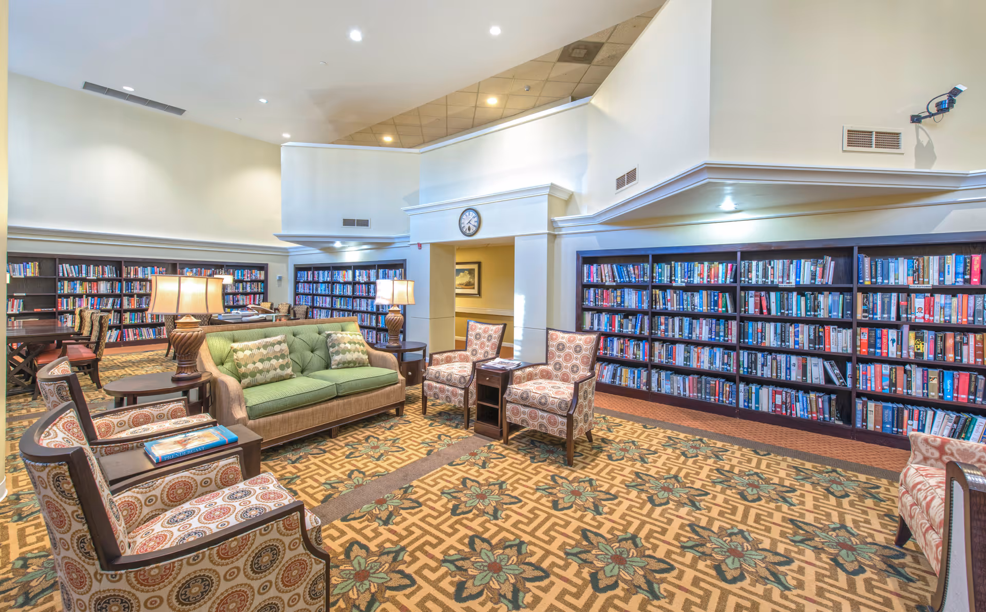Spacious lounge-style library with bookshelves lining the walls, upholstered chairs, a green sofa, and table lamps.
