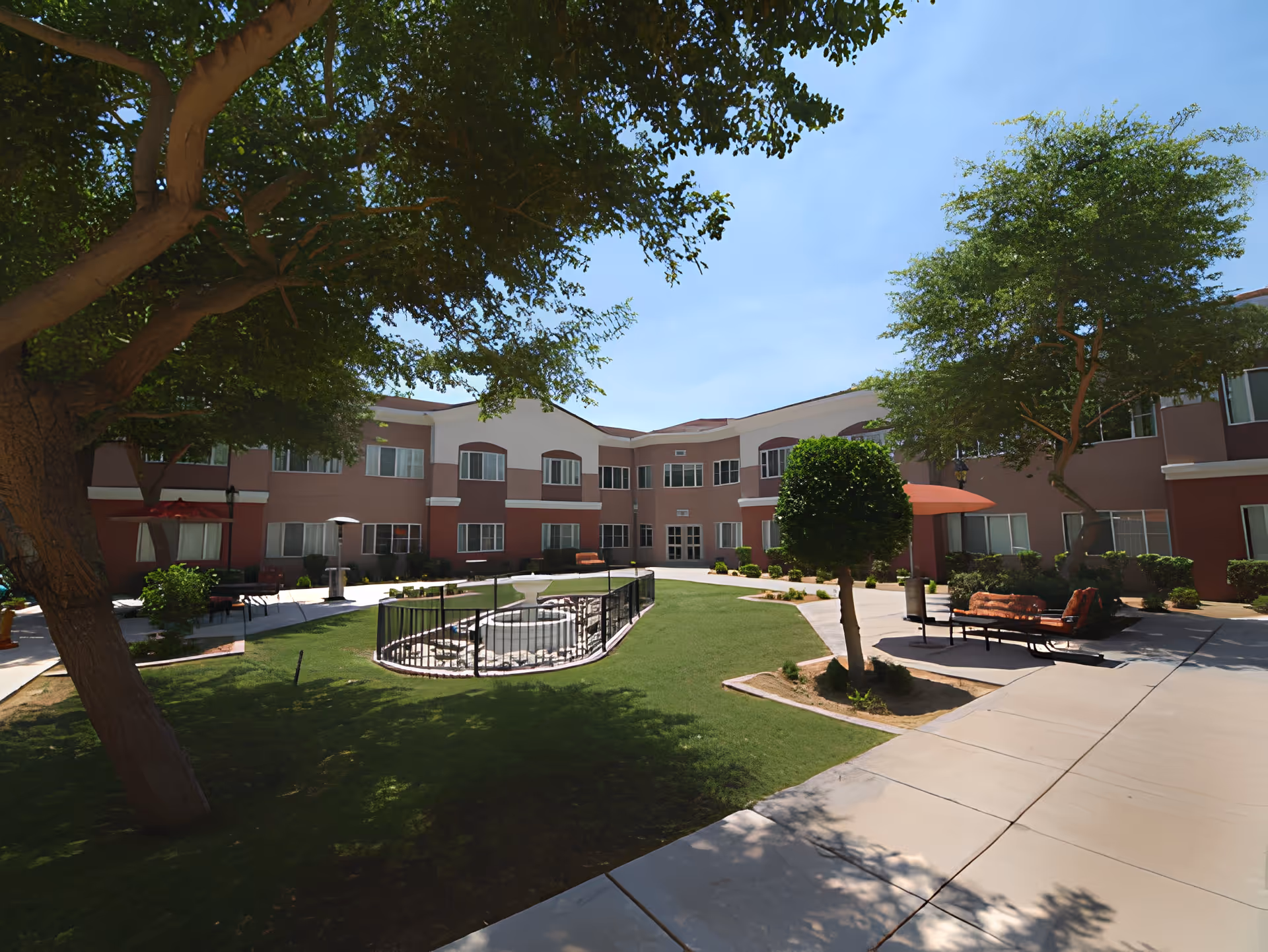 Outdoor courtyard area of Chaparral Winds Assisted Living featuring a central fountain surrounded by a black metal fence, green grass, trees providing shade, and seating areas with benches and umbrellas. The two-story building with multiple windows encloses the courtyard.