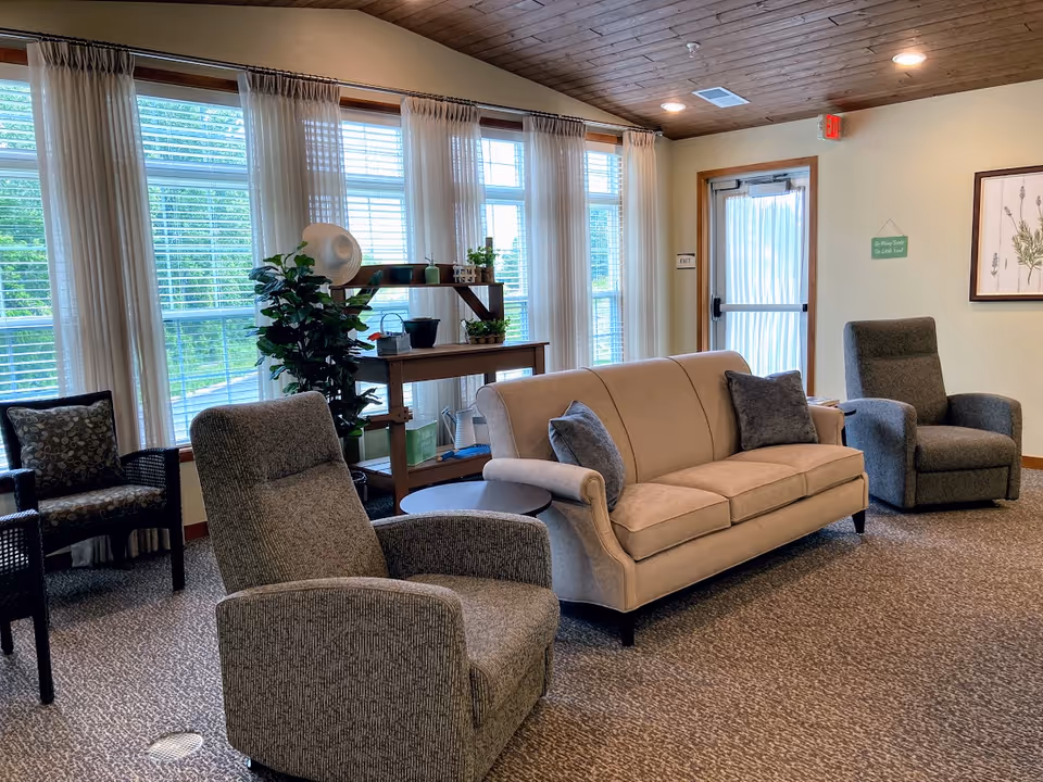 A cozy senior living common area with a beige sofa, two gray armchairs, and a black chair with patterned cushions. Large windows with sheer white curtains allow natural light to fill the room. A wooden shelf with potted plants and decorative items is placed near the windows. The room has a carpeted floor and a wooden ceiling with recessed lighting. A door with a sheer curtain is visible on the right side of the room.