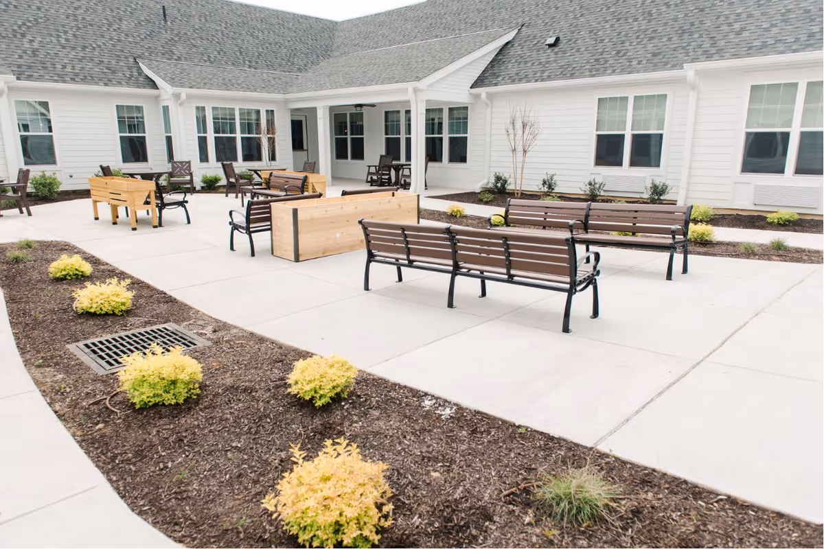 Outdoor courtyard area of a senior living facility with several wooden benches and planter boxes arranged on a concrete patio. The building surrounding the courtyard has white siding and multiple windows. Small shrubs and plants are planted in mulched beds around the patio.