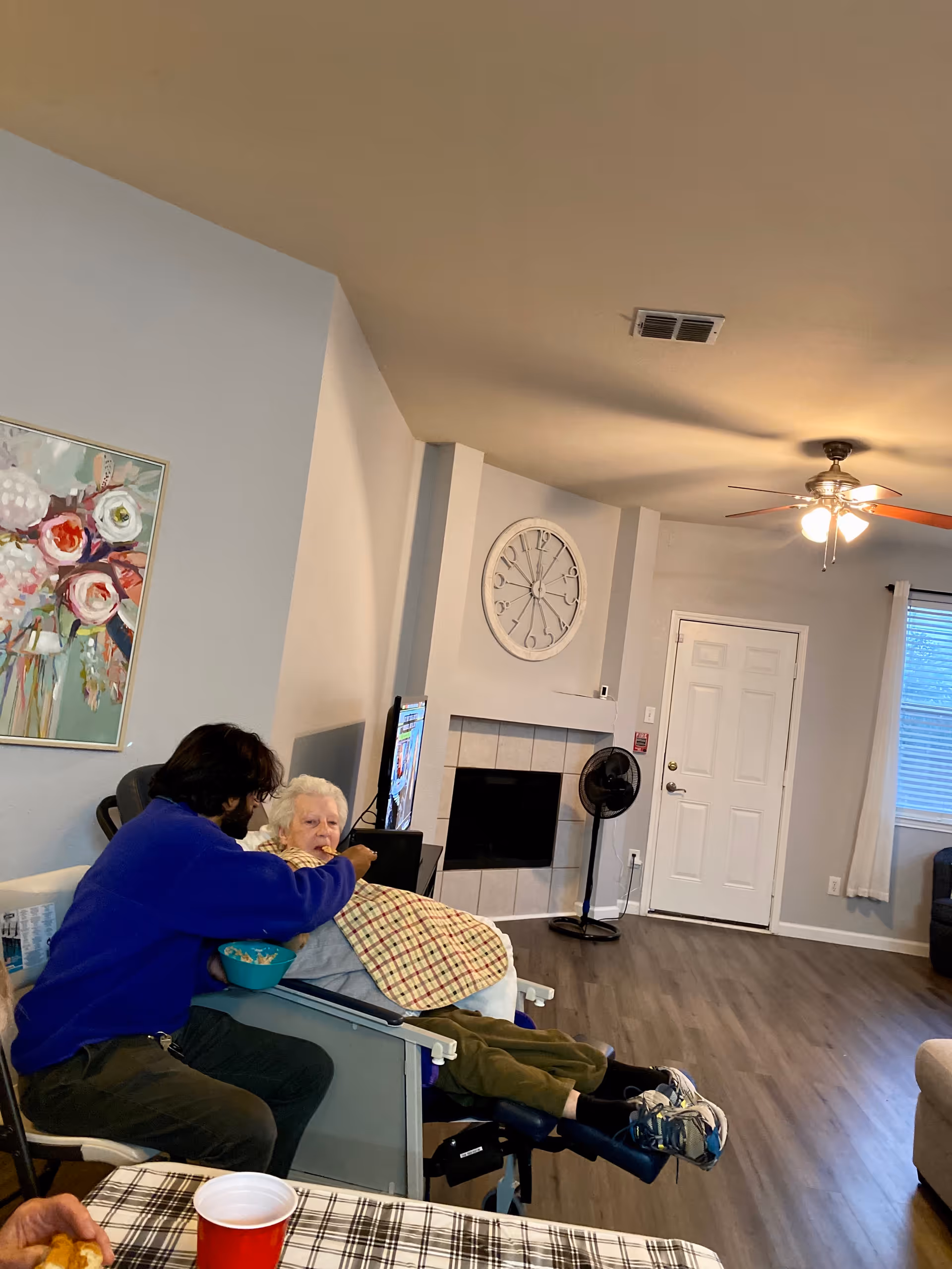 A man in a blue sweater is feeding an elderly woman who is seated in a reclining chair with a checkered blanket over her lap. They are in a living room with a large clock on the wall above a fireplace, a standing fan, a ceiling fan with lights, and a window with white curtains. A colorful floral painting is visible on the wall, and a table with a red cup and a checkered tablecloth is in the foreground.