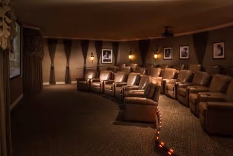 A cozy home theater room with multiple rows of brown leather recliner chairs arranged in a curved layout facing a large screen. The room has dim lighting with wall sconces and framed black-and-white photographs on the walls. Dark curtains cover the windows, creating a warm and inviting atmosphere.