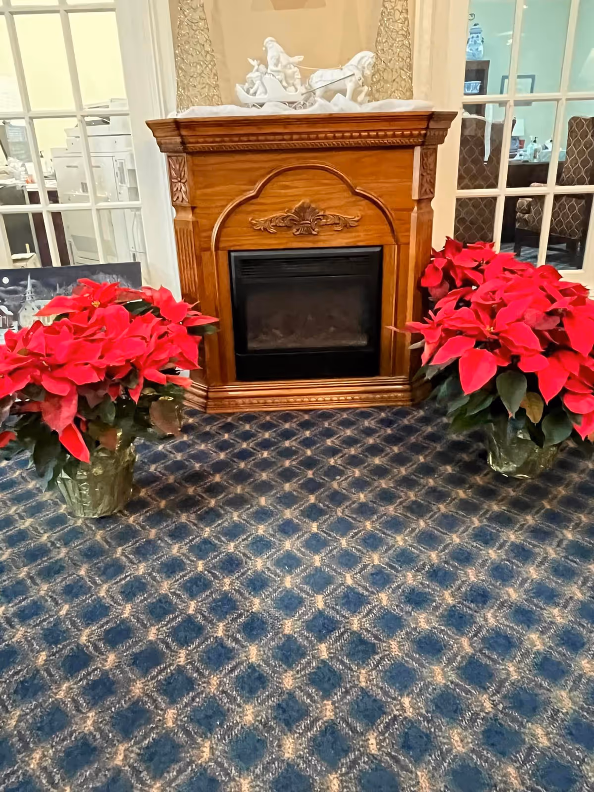 A wooden electric fireplace with ornate carvings is centered against a wall with glass-paneled doors on either side. Two large pots of vibrant red poinsettia plants are placed symmetrically on the blue patterned carpet in front of the fireplace. A white decorative sculpture is displayed on the mantel above the fireplace.