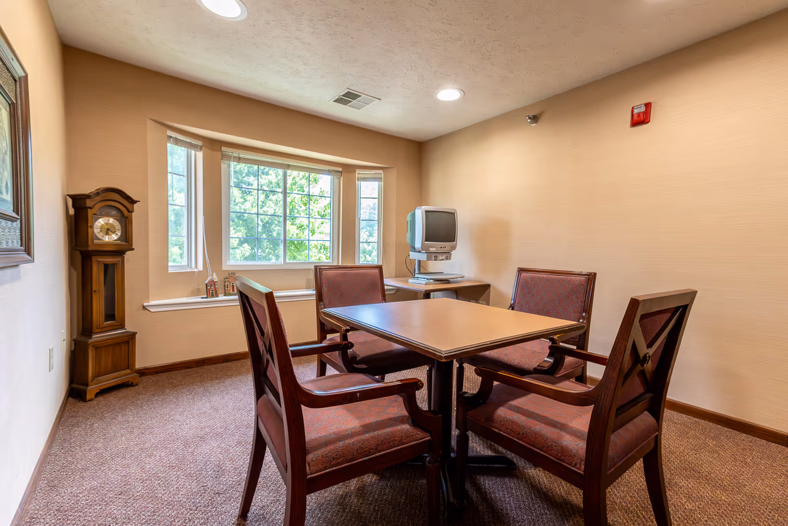 A small room with a square wooden table surrounded by four upholstered chairs. There is a large window with a view of green trees, a wooden grandfather clock in the corner, and an old-style CRT television on a small table against the wall. The room has beige walls and carpeted floor.