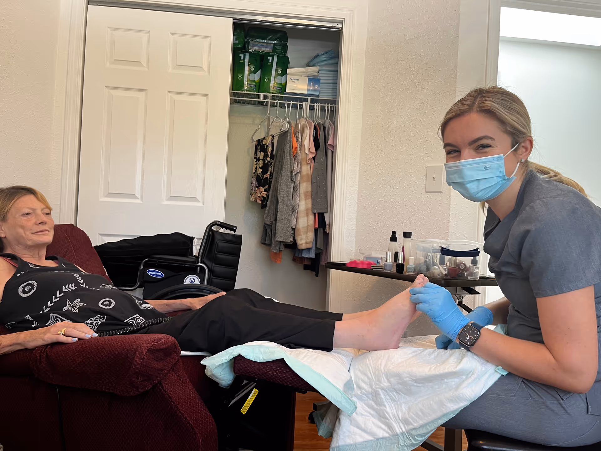 A woman wearing a medical mask and gloves is providing foot care to an elderly woman who is seated in a maroon recliner chair. The elderly woman has her legs extended and resting on a padded surface. Behind them, there is an open closet with clothes hanging and medical supplies on the shelf. A wheelchair is also visible in the background.
