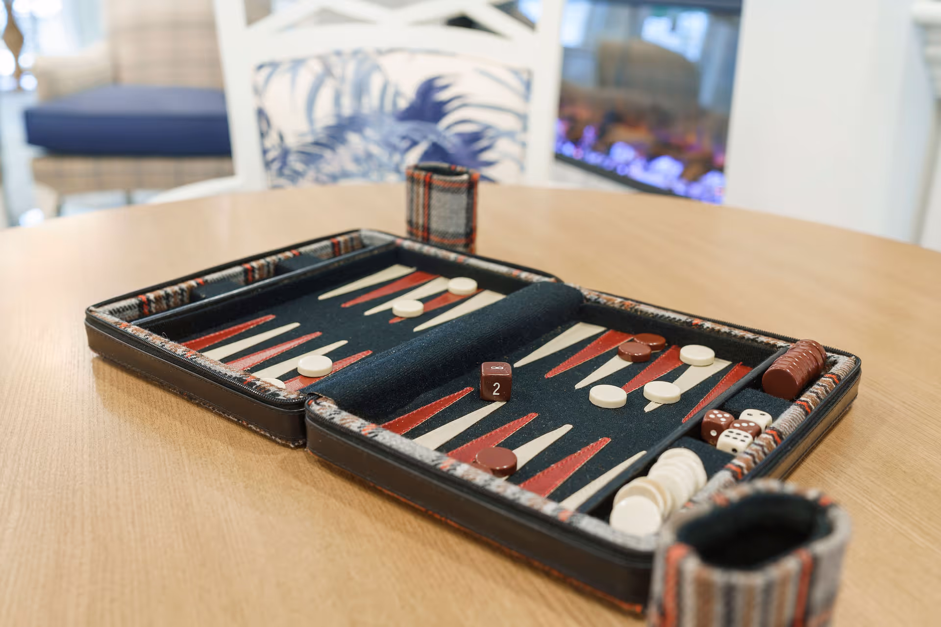 A backgammon board set up on a wooden table with checkers and dice, with a cozy chair and a fireplace in the background.