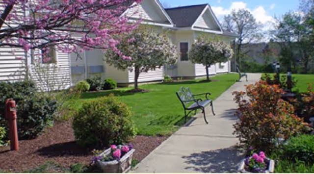 A landscaped outdoor walkway beside a light-colored building with blooming trees, green grass, benches, and flower beds under a blue sky with some clouds.