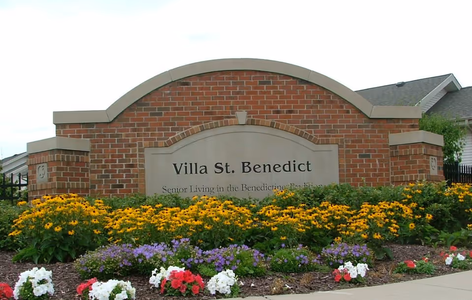 A brick sign for Villa St. Benedict surrounded by colorful flowers including yellow, purple, white, and red blooms, with a sidewalk in front and residential buildings partially visible in the background.