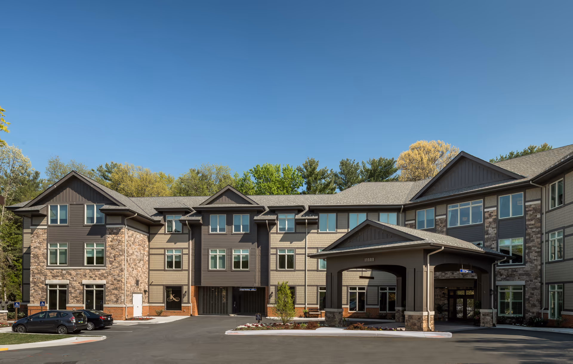 Front exterior of a three-story senior living facility with a covered entrance canopy, parking spaces, and landscaped grounds under a clear blue sky.