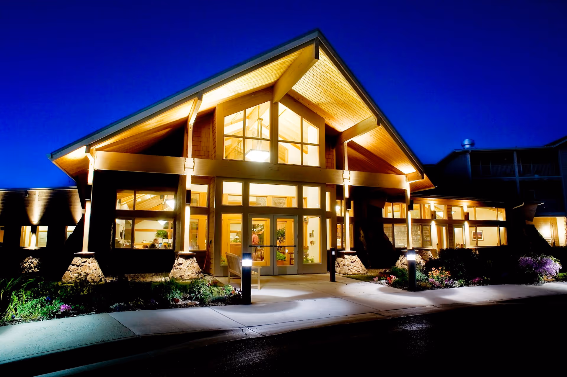 Exterior nighttime view of Bozeman Health Hillcrest Senior Living, Aspen Pointe building entrance with warm interior lights shining through large windows and a peaked roof structure, surrounded by landscaped plants and a sidewalk.