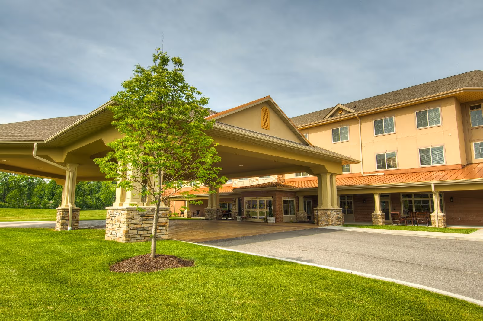 Exterior view of Garden Plaza of Florissant showing the main entrance with a covered driveway supported by stone pillars, a small tree in the foreground, well-maintained green lawn, and a multi-story building with windows in the background under a partly cloudy sky.