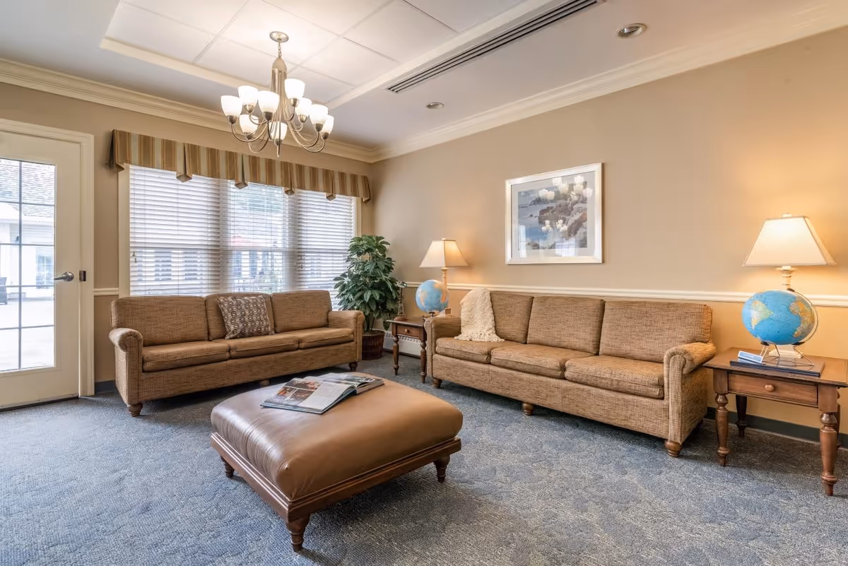 A cozy living room with two brown upholstered sofas facing each other, a large brown leather ottoman in the center with an open magazine on it, a side table with a globe and lamp, a potted plant near the window, and a framed picture on the beige wall. The room has a large window with blinds and a striped valance, a chandelier hanging from the ceiling, and carpeted floor.