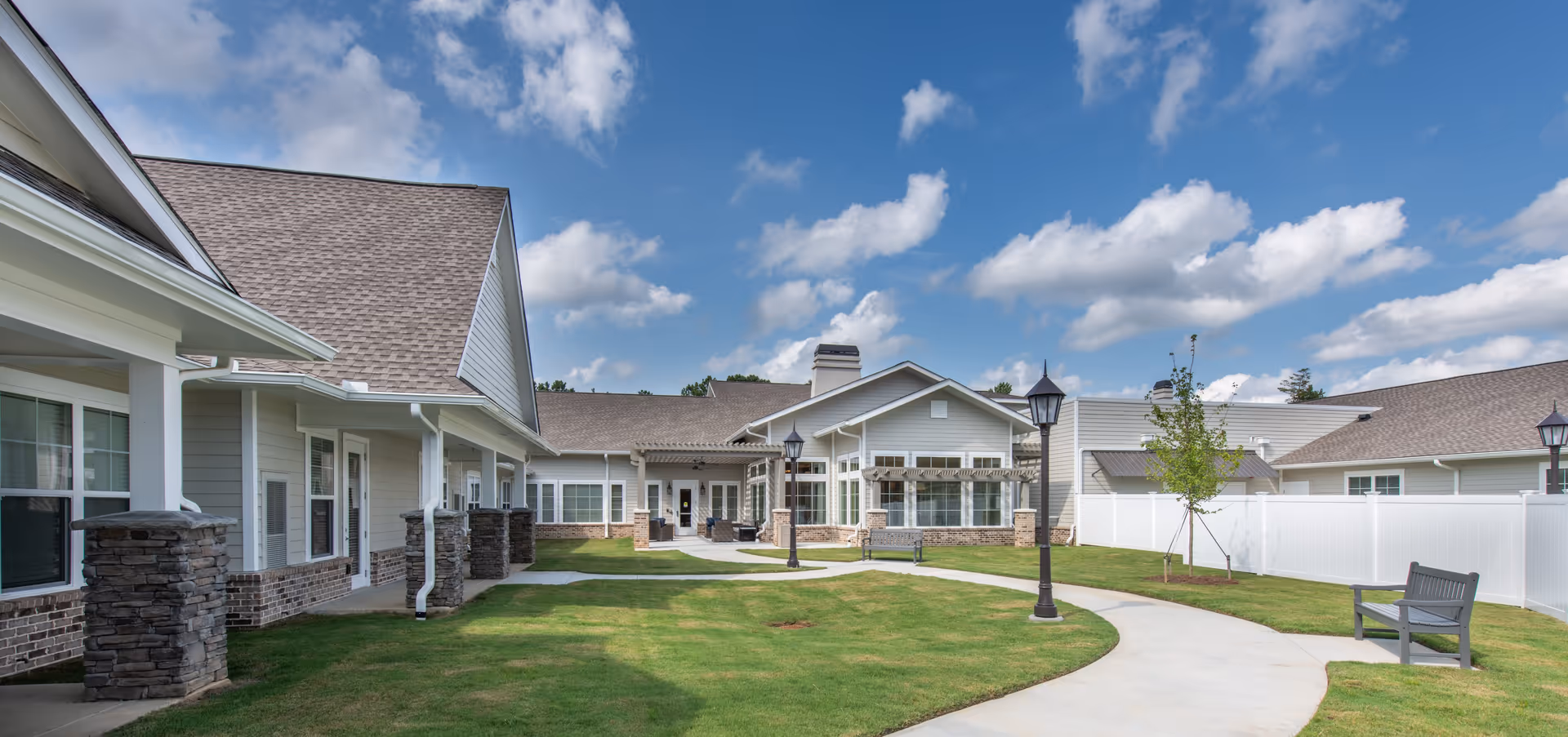 Outdoor view of a senior living facility with a curved concrete pathway, green lawn, benches, lampposts, and single-story buildings with light-colored siding and stone accents under a partly cloudy blue sky.