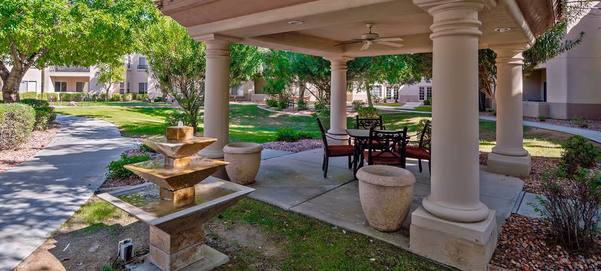 Outdoor covered seating area with a round table and four chairs with red cushions, surrounded by large white columns. In front of the seating area is a tiered stone water fountain. The area is part of a landscaped garden with green grass, trees, bushes, and a paved walkway leading through the garden. Residential buildings are visible in the background.