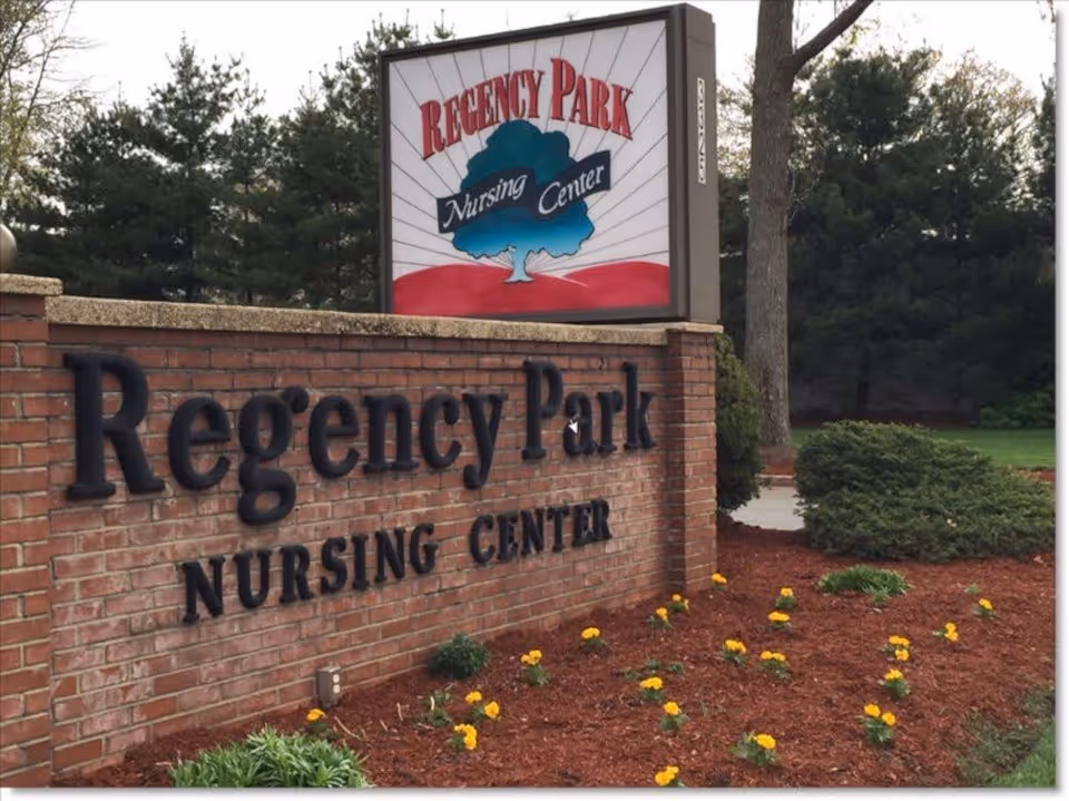 Outdoor view of a brick sign for Regency Park Nursing Center with a landscaped area featuring small yellow flowers and green shrubs, and trees in the background.