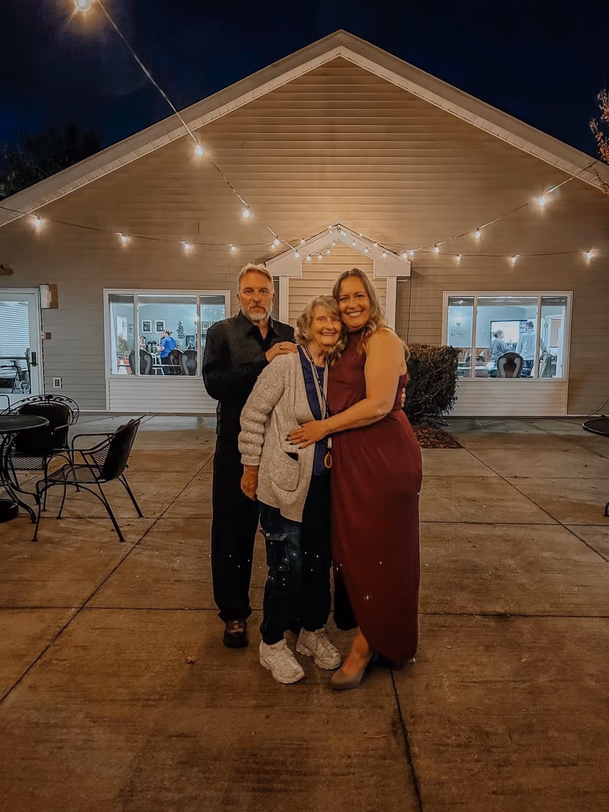 Three people standing outside a building at night, with string lights overhead. The group consists of an elderly woman in a gray cardigan and sneakers, a man in a black shirt, and a woman in a sleeveless maroon dress. The building behind them has large windows showing people inside and a small porch with a peaked roof.