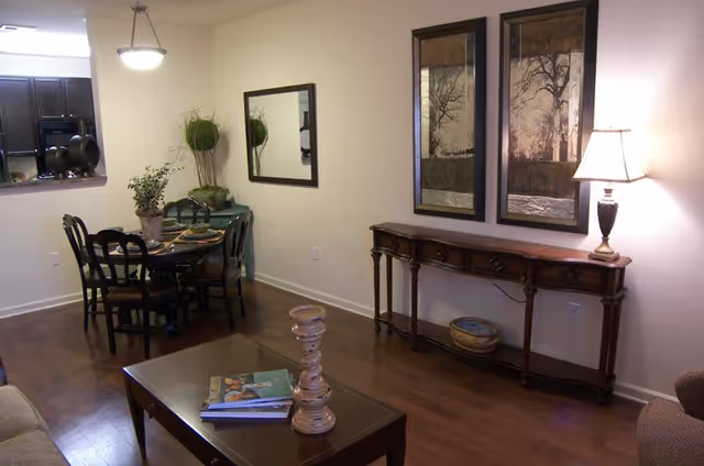 A cozy living and dining area with wooden flooring. The dining area has a round table with four chairs and a plant centerpiece. A large mirror hangs on the wall above a small green cabinet with decorative plants. The living area features a wooden coffee table with books and a candle holder, a long wooden console table with a lamp and decorative bowl, and two framed artworks on the wall.