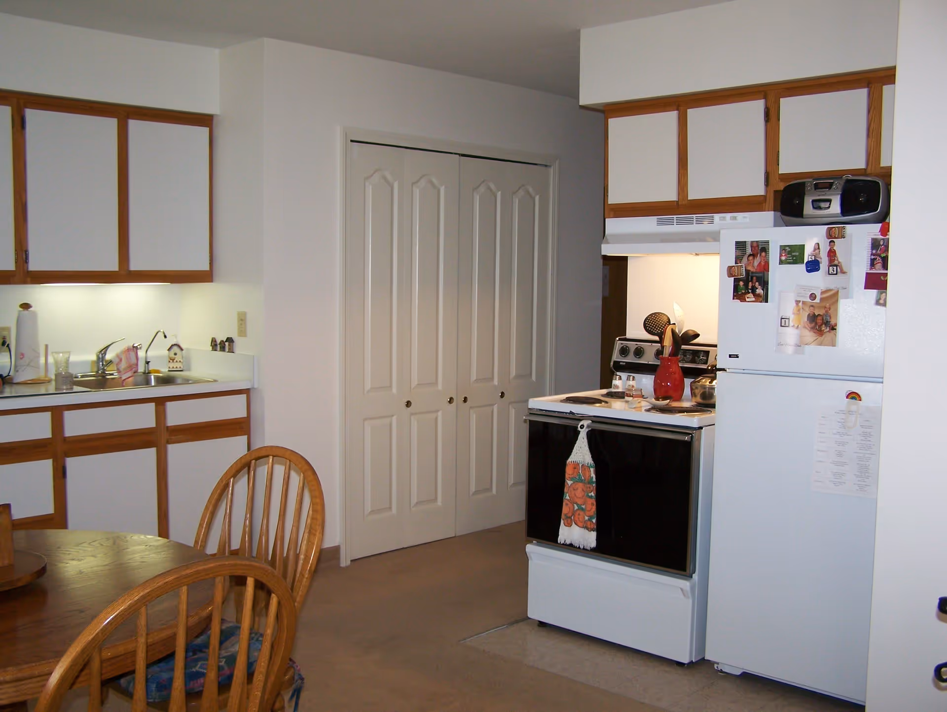 A kitchen area in an independent living facility featuring wooden cabinets with white doors, a white refrigerator with magnets and photos, a white stove with a red vase and kitchen utensils on top, a double sink with a faucet, and a wooden dining table with chairs in the foreground.