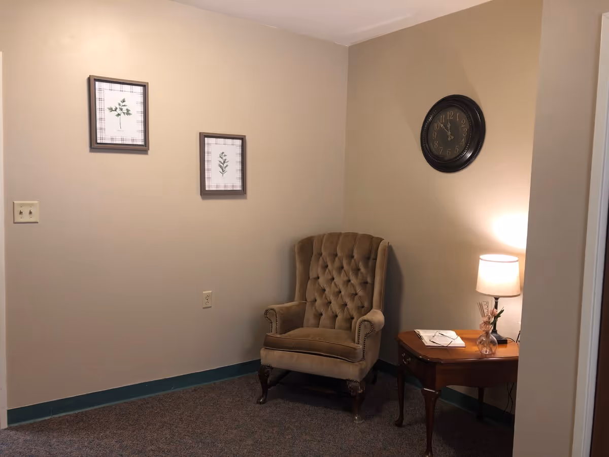 A cozy corner of a room with a brown tufted armchair, a wooden side table with a lamp, a book, and a small vase with flowers. Two framed botanical prints hang on the beige wall, and a black wall clock shows the time as 11:55.