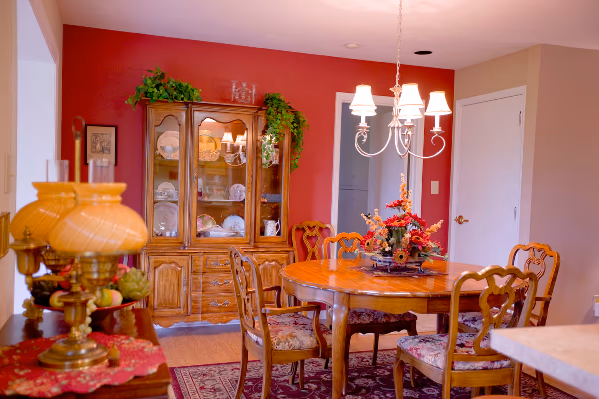 A traditional dining room with a wooden dining table and four upholstered chairs. A floral centerpiece is on the table. Behind the table is a wooden china cabinet displaying plates and glassware, with green plants on top. The walls are painted red and beige, and a chandelier with five lampshades hangs above the table. A vintage-style lamp is visible on the left side of the image.