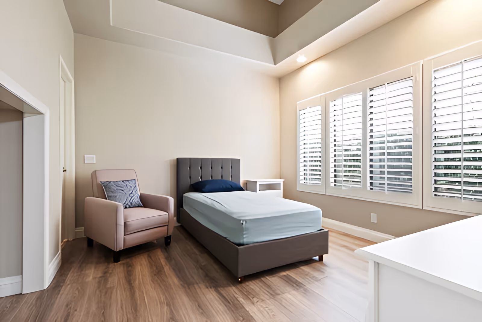 A bright and clean bedroom with a single bed featuring a gray upholstered headboard and light blue bedding. Next to the bed is a white nightstand, and a comfortable pink armchair with a decorative pillow is placed nearby. The room has large windows with white plantation shutters allowing natural light to fill the space, and the floor is covered with wood-style flooring.