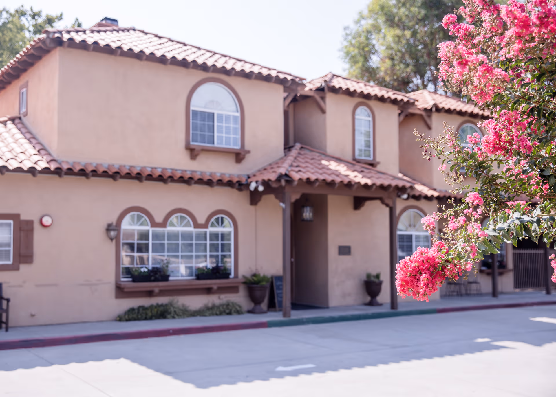 Exterior view of a two-story building with tan stucco walls and red tile roofing, featuring arched windows with brown trim and flower boxes. Pink flowering tree branches are visible in the foreground on the right side.