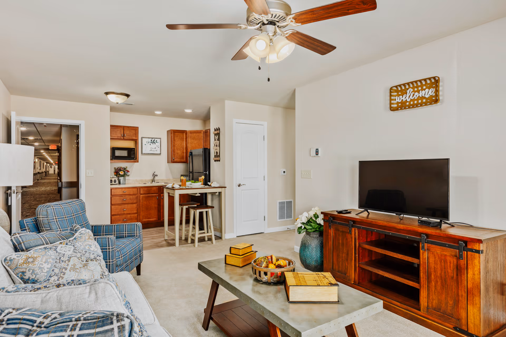 A cozy living area in an assisted living facility featuring a plaid armchair and a sofa with patterned pillows, a wooden coffee table with decorative items, a flat-screen TV on a wooden TV stand, and a small kitchen area with wooden cabinets, a refrigerator, microwave, and a small dining table with two stools. A ceiling fan with lights is mounted on the ceiling, and a 'welcome' sign is hung on the wall above the TV.