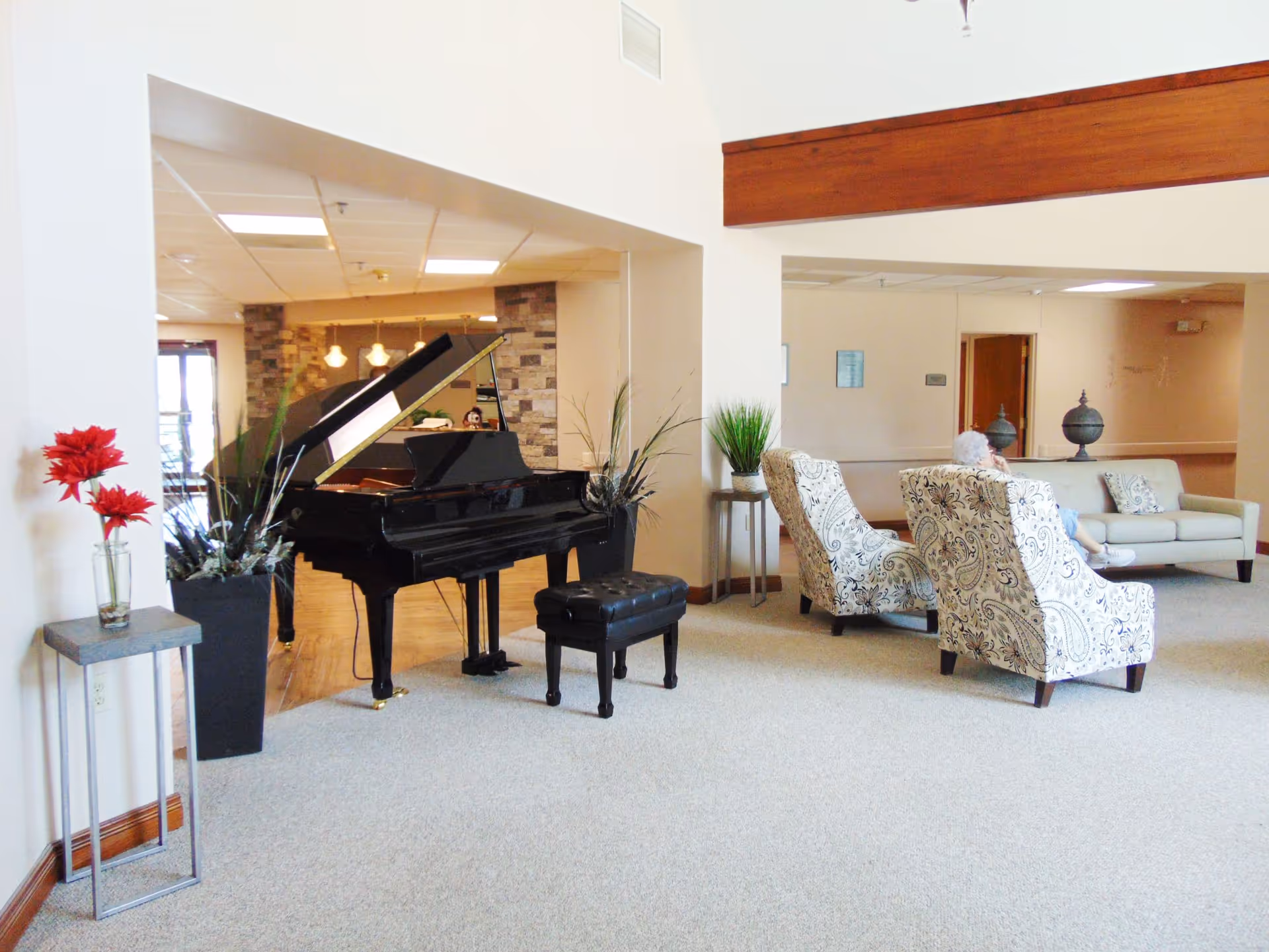 Bright senior living common room with a black grand piano, patterned armchairs, a sofa, and decorative plants.