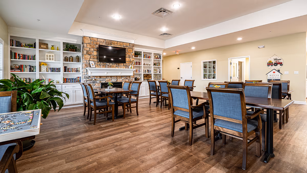 A spacious common area with multiple wooden tables and blue upholstered chairs arranged neatly on a wood floor. The room features a stone fireplace with a mounted flat-screen TV above it, flanked by white built-in bookshelves filled with books and decorative items. There are plants and a puzzle table visible in the foreground, and the walls are painted a light beige color with recessed lighting overhead.