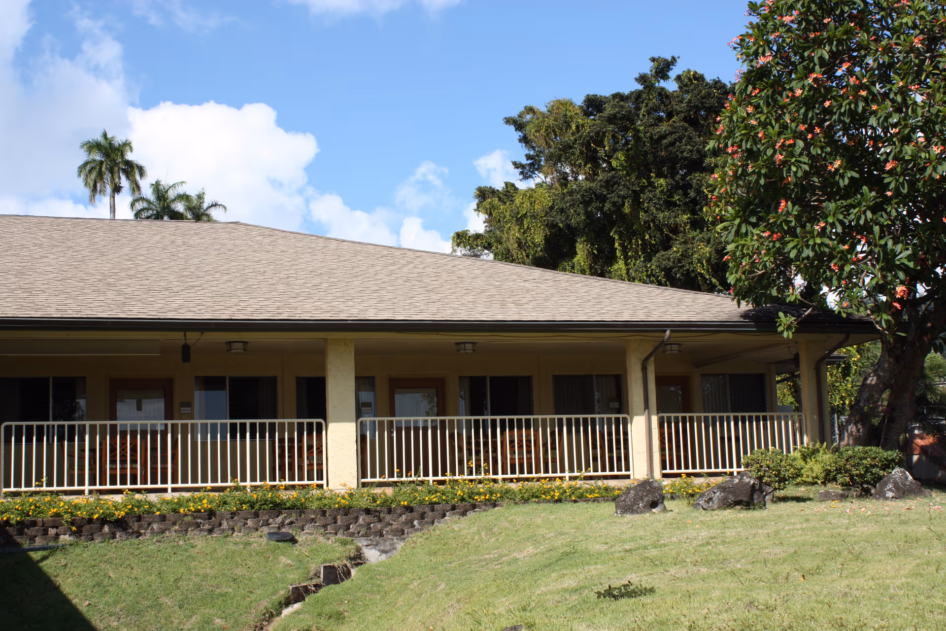 Exterior view of a single-story building with a covered porch and railing, surrounded by green grass, trees, and a partly cloudy blue sky.