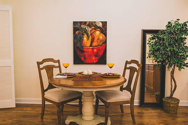 Round wooden dining table set for two with chairs, a fruit painting on the wall, and a potted plant beside a mirror.