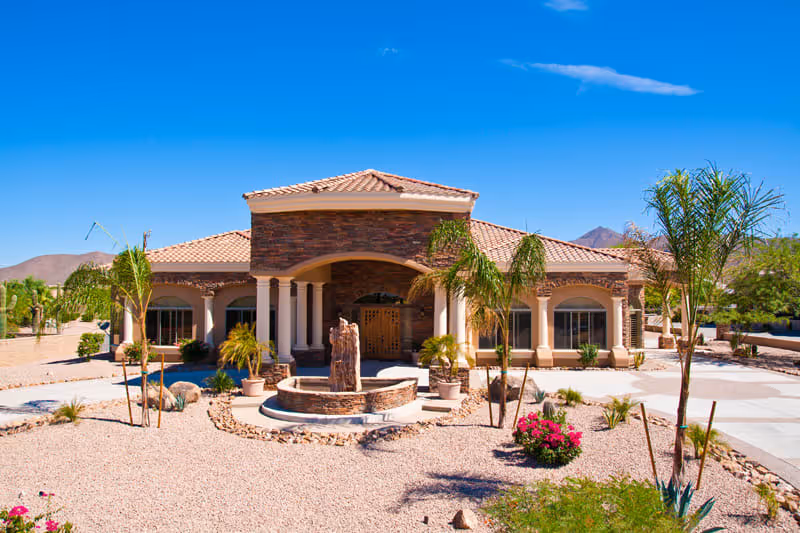 Front exterior view of a single-story building with a tiled roof and stone facade, surrounded by desert landscaping including palm trees, rocks, and flowering plants under a clear blue sky.