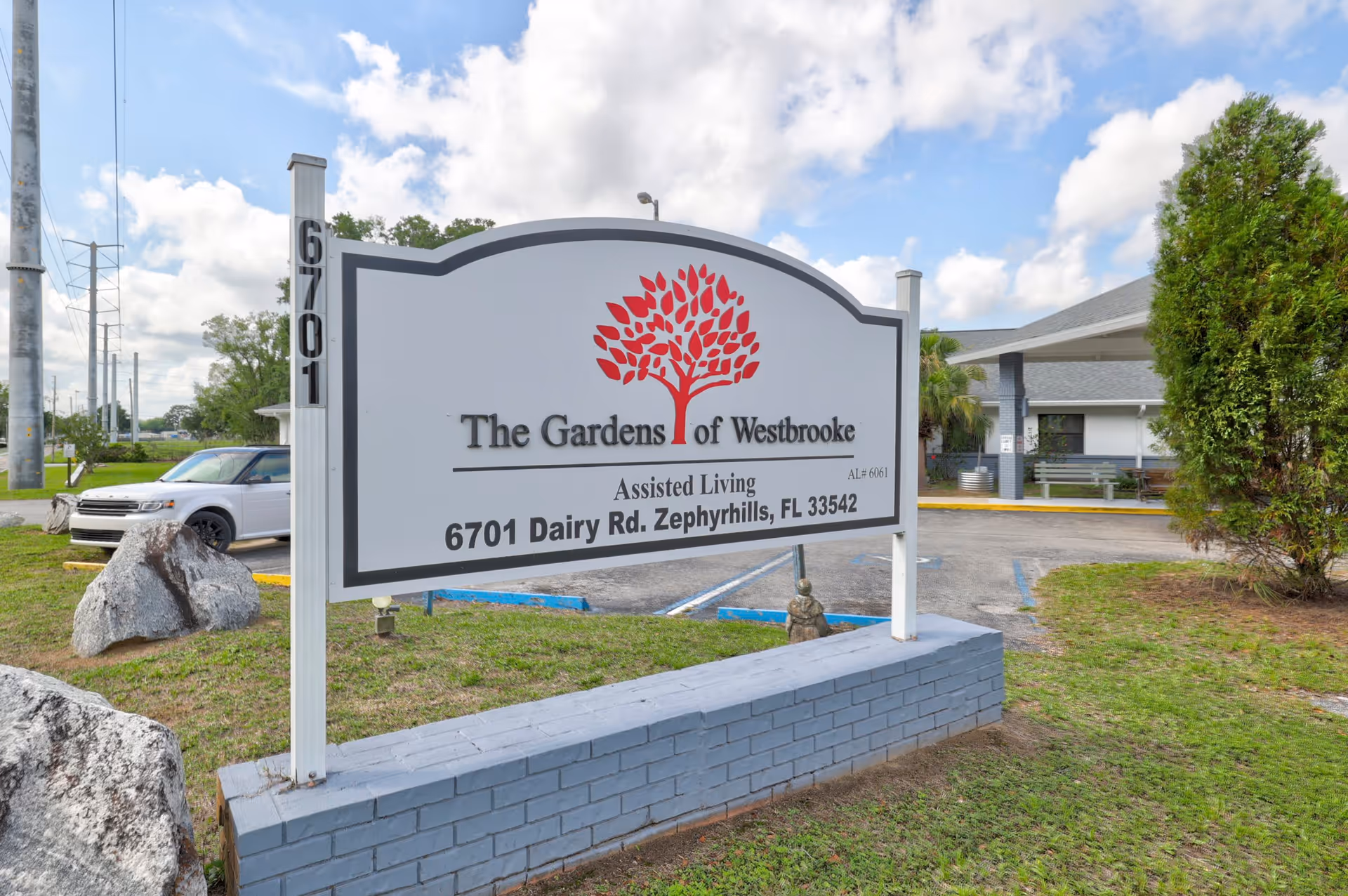 Outdoor view of the entrance sign for The Gardens of Westbrooke assisted living facility located at 6701 Dairy Rd, Zephyrhills, FL 33542, with a building and parking area in the background under a partly cloudy sky.