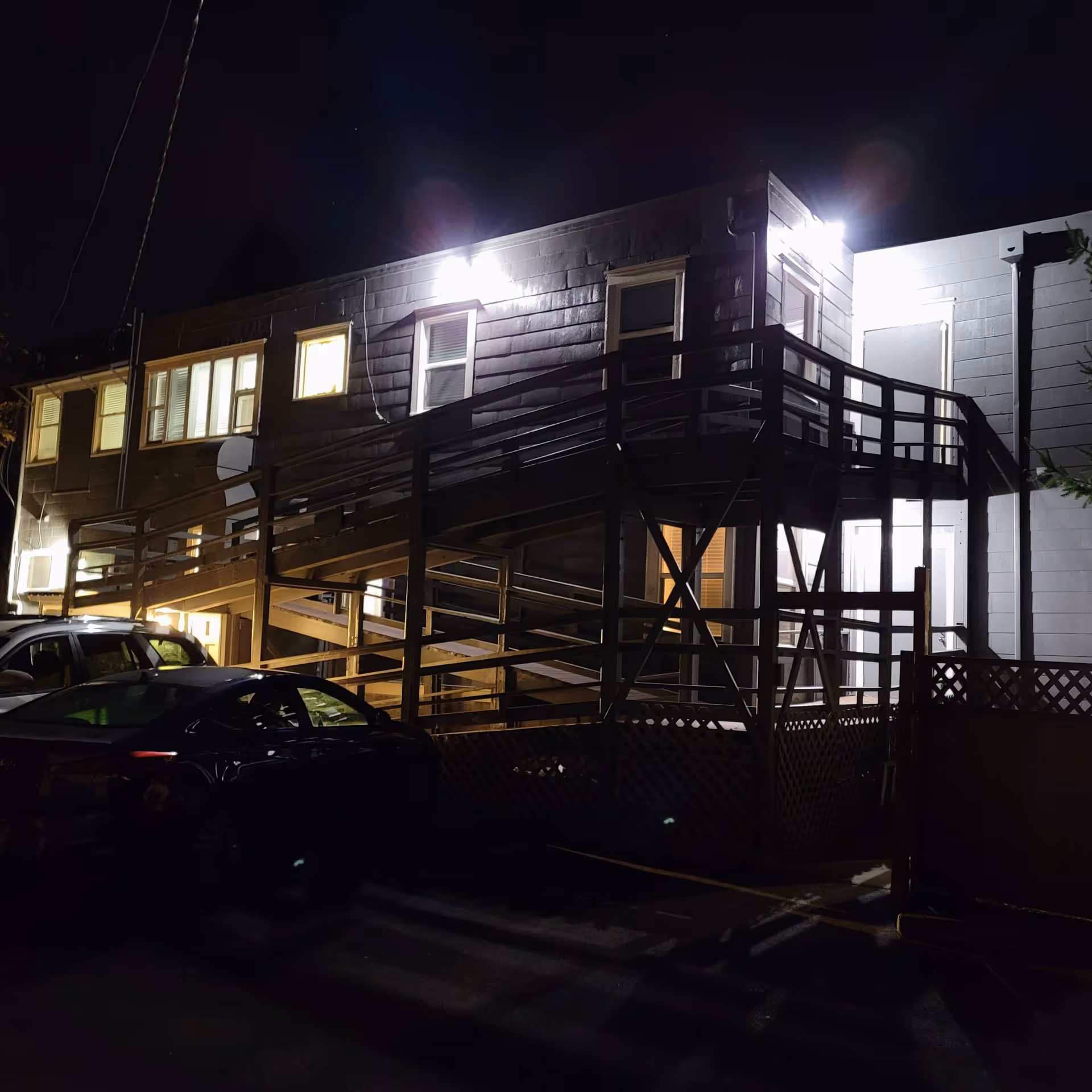 Nighttime exterior view of a two-story building with illuminated windows and outdoor lights. A wooden ramp with railings leads to the second floor. Several parked cars are visible in the foreground.
