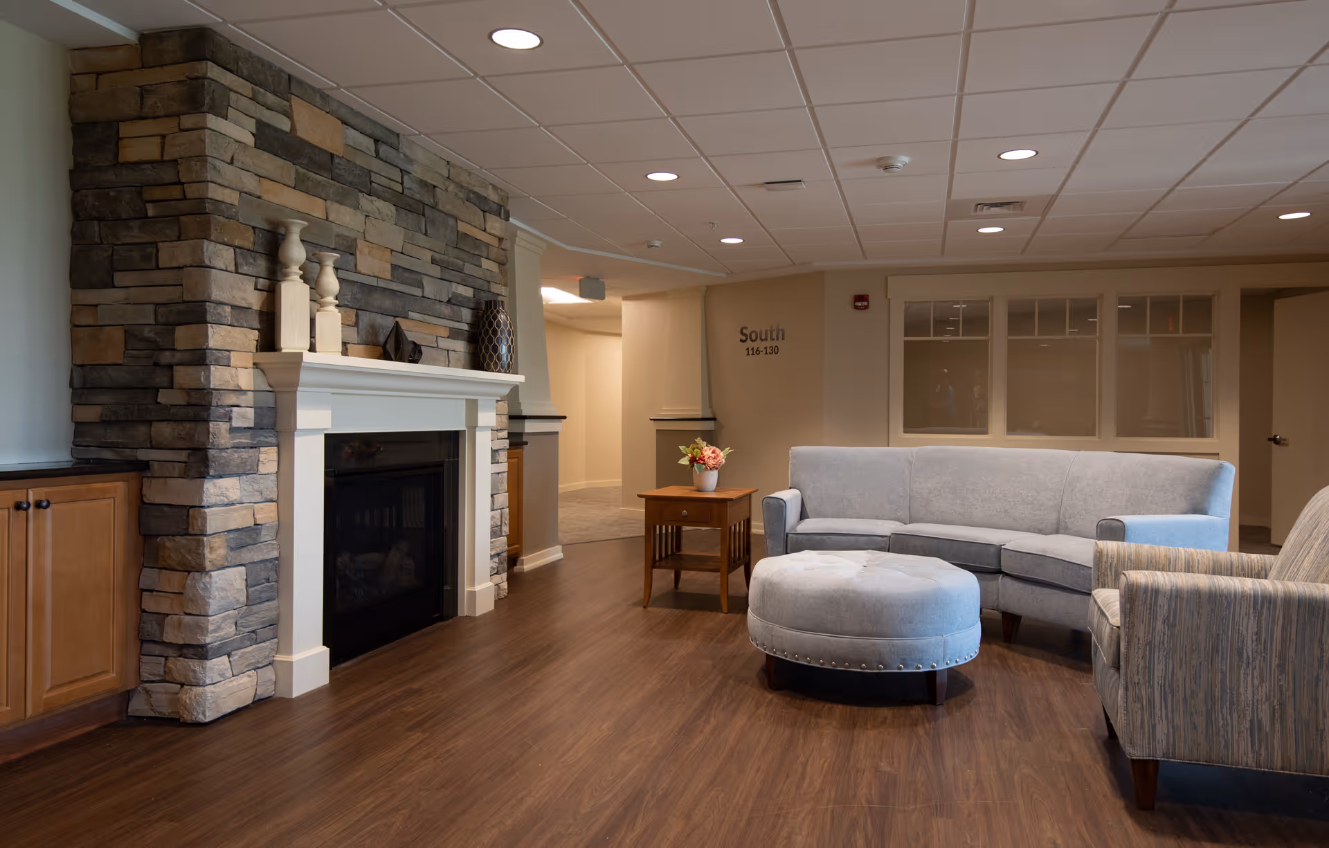 A cozy living room area in a senior living facility featuring a stone fireplace with a white mantel, a light gray sectional sofa, a round upholstered ottoman, a wooden side table with a flower vase, and a comfortable armchair. The room has wood flooring and a ceiling with recessed lighting. A hallway labeled 'South 116-130' is visible in the background.