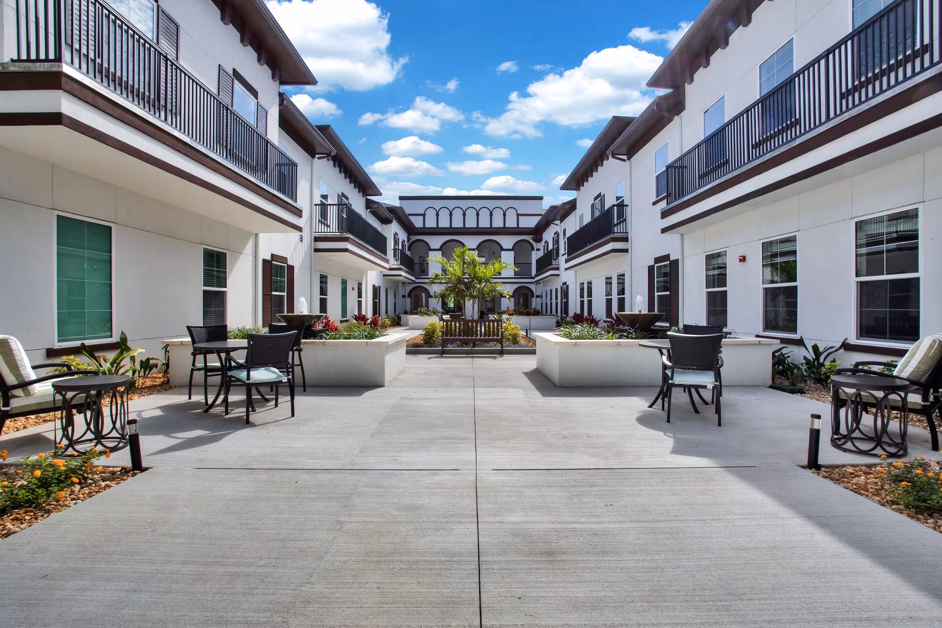 Outdoor courtyard area of The Blake at LPGA featuring a spacious concrete patio with several black metal tables and chairs, cushioned seating, large planters with greenery and flowers, a central bench, and a two-story building with balconies and windows surrounding the courtyard under a blue sky with scattered clouds.
