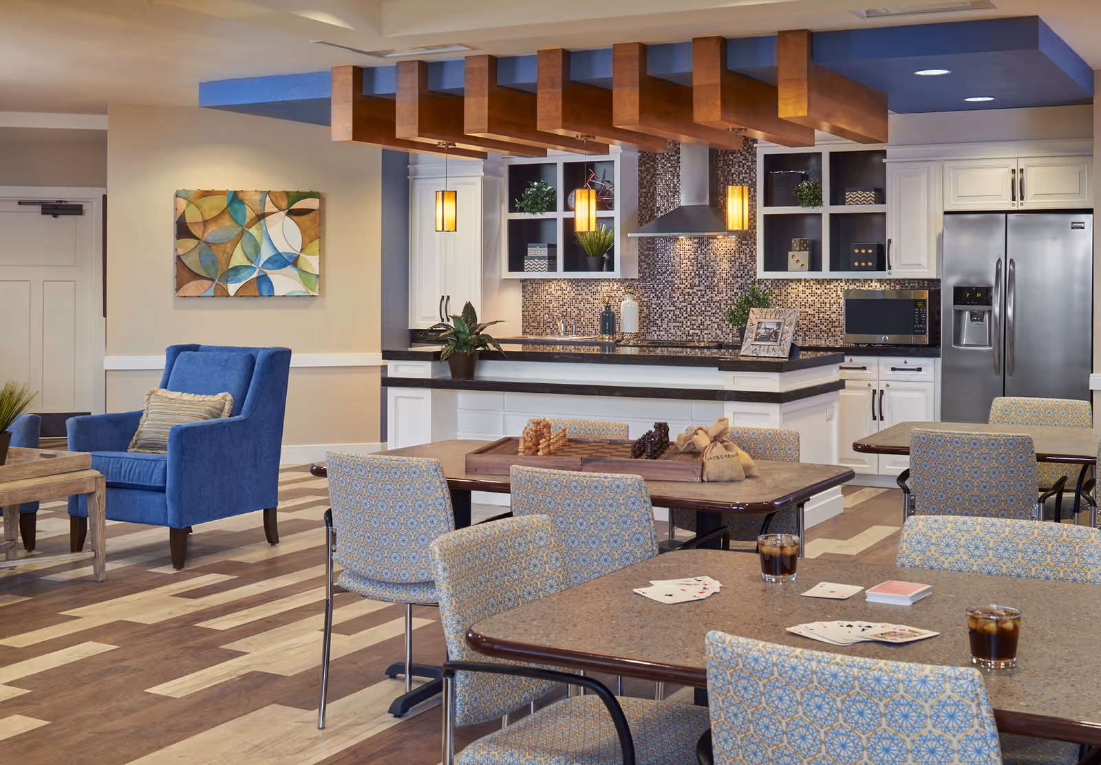 A bright communal dining area with tables and patterned chairs, a kitchen island with pendant lights, and blue lounge chairs in the background.