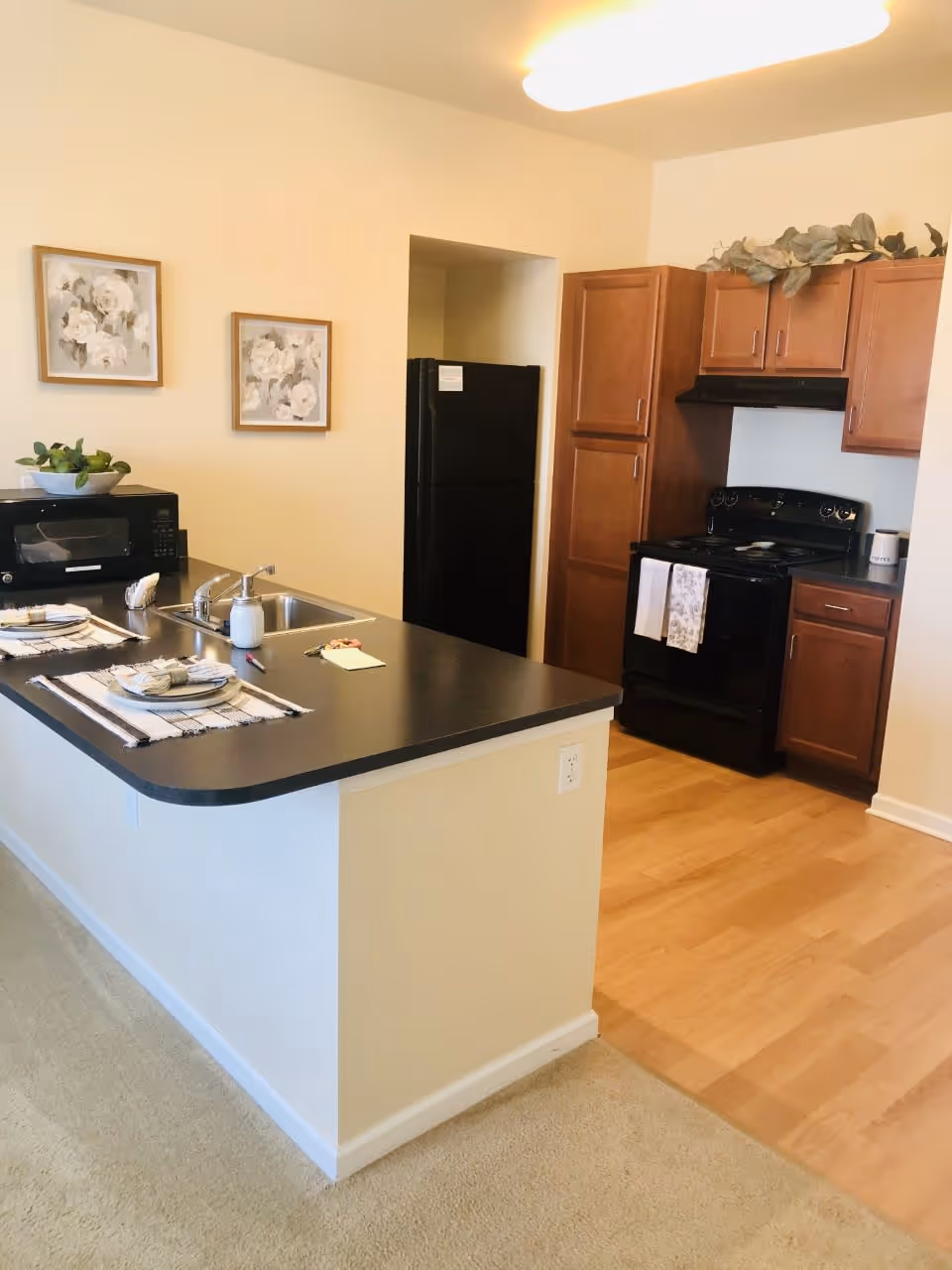 A kitchen area featuring a black countertop island with a sink, a microwave, and place settings. The kitchen has wooden cabinets, a black refrigerator, and a black stove with towels hanging on the handle. Two framed floral artworks hang on the wall above the microwave.