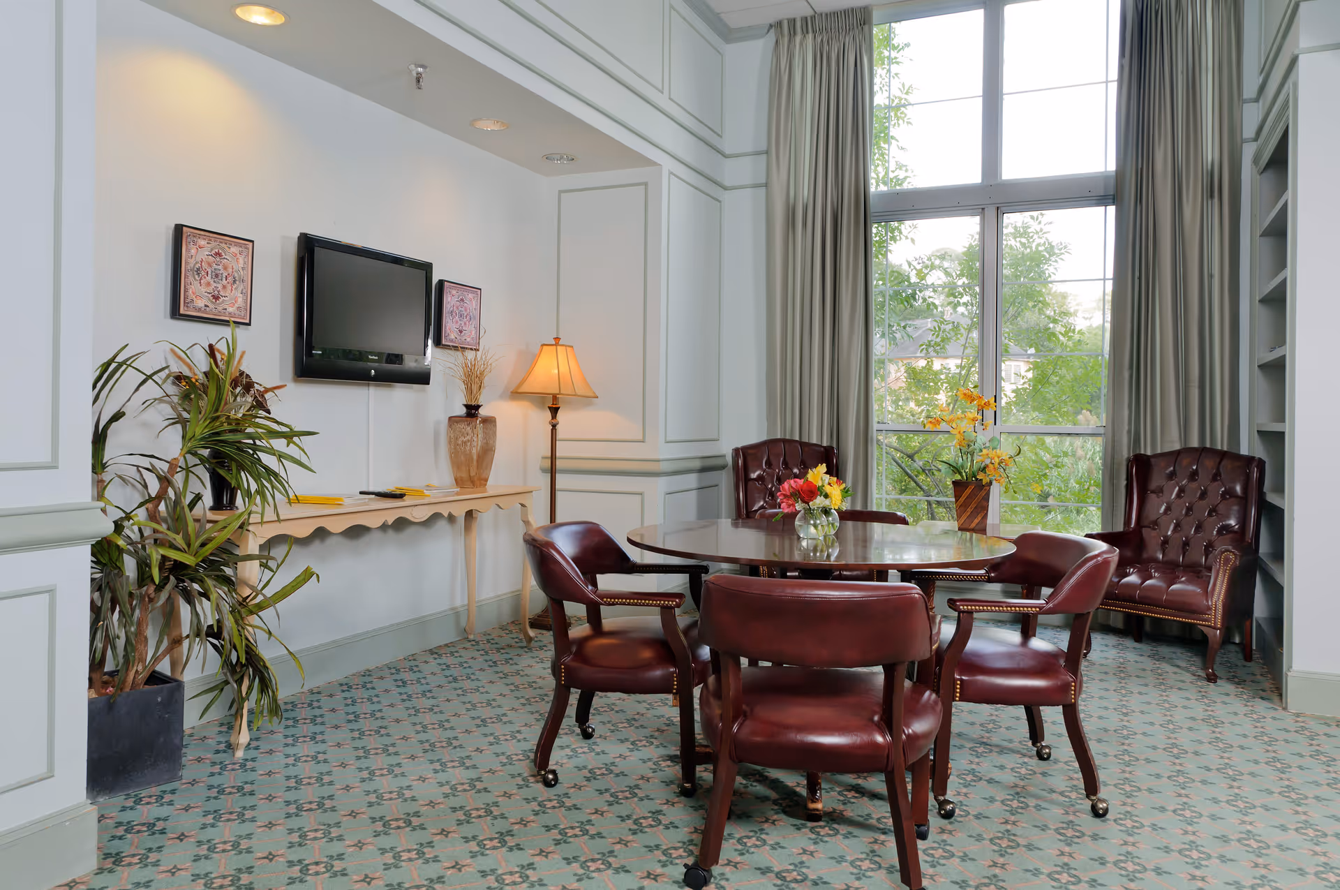 A cozy sitting area with a round wooden table surrounded by four maroon leather chairs. Two matching leather armchairs are placed near a large window with long beige curtains, allowing natural light to brighten the room. A tall floor lamp and a decorative vase with flowers sit on a console table against the wall, which also features a mounted flat-screen TV and two framed artworks. A potted plant is positioned in the corner, and the floor is covered with a patterned carpet.