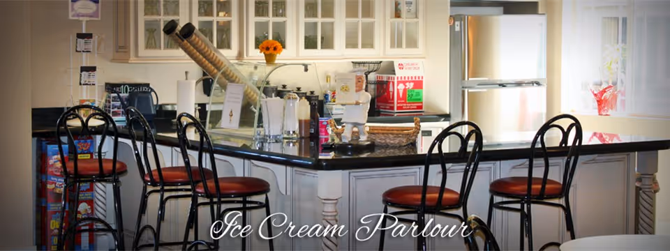 Interior view of an ice cream parlour with a black countertop island surrounded by six black metal chairs with red seats. Behind the counter are white cabinets with glass doors, a refrigerator, and various items including ice cream cones and condiments.